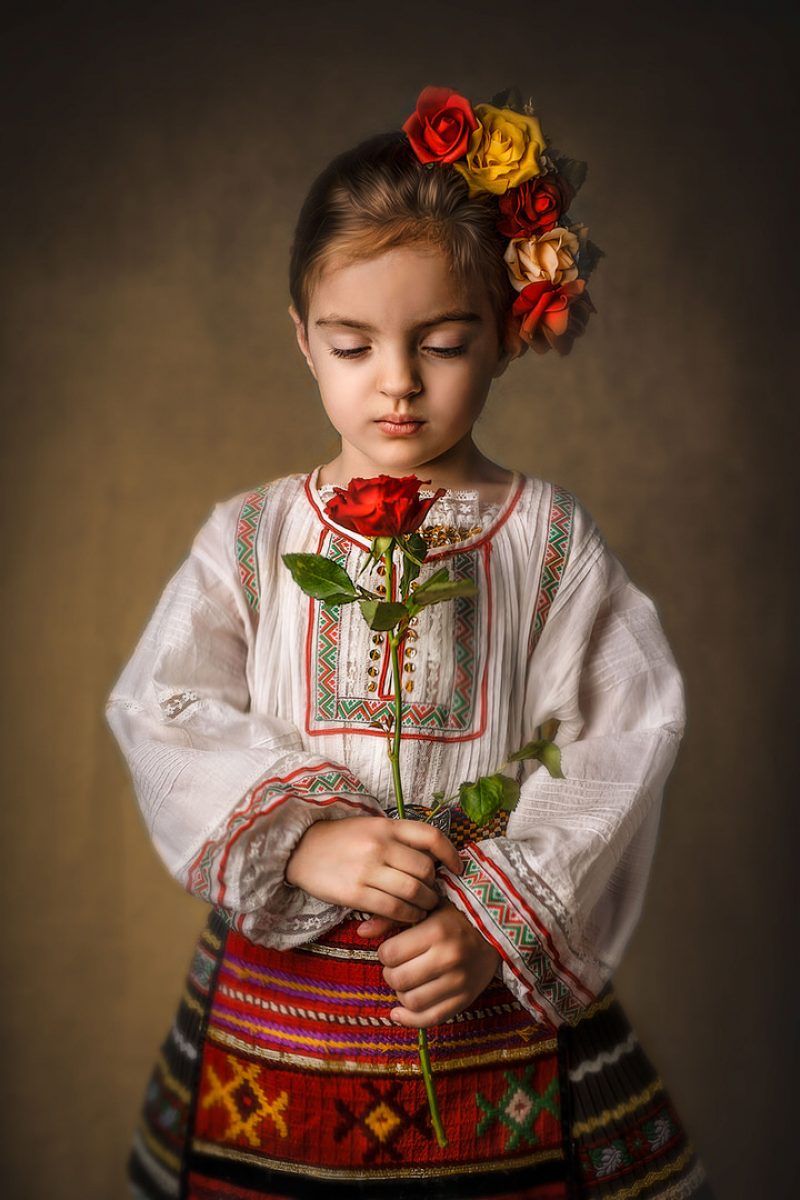 Fine art portrait — child in traditional dress holding a red rose by Iva Kostova